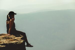 A woman enjoys a calm moment sitting on a cliff's edge against a foggy mountainous backdrop.