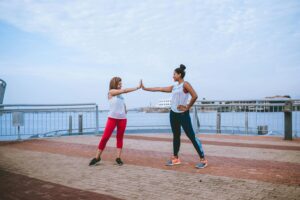 Two women high-fiving while exercising outdoors near the waterfront, promoting fitness and teamwork.